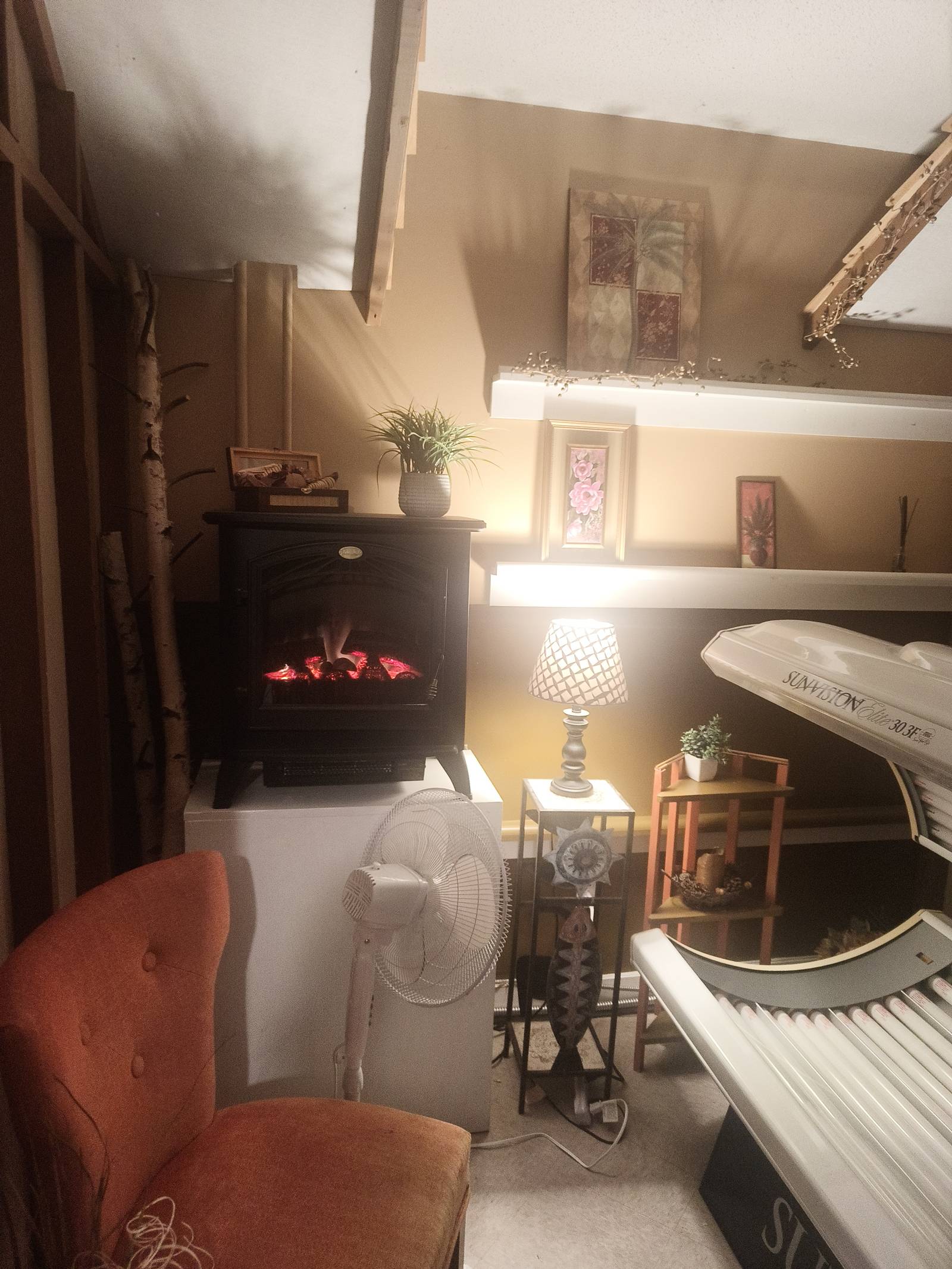 Private tanning room at Tanorama Falmouth with a SunVision Pro 30/3E tanning bed, an electric fireplace, a lamp, a fan, and an armchair